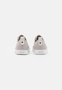 Beiga canvas sneakers med vita gummisulor, med en baksida med en knappdetalj och logotypen "JOOP!" präglad på sidan.