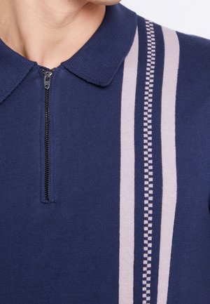 Granatowy dzianinowy polo z suwakiem z przodu i dwoma pionowymi beżowymi paskami, z których jeden ma wzór szachownicy, po prawej stronie.