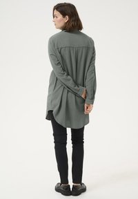 Pikad varrukad, oliivrohelise särgi ümardatud äärega, pehme tekstuuriga ja tagumise õla detailiga, kombineeritud mustade kitsaste teksade ja massiivsete mustade kingadega.