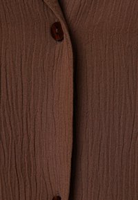 Tessuto marrone con texture e ripples verticali, dotato di chiusura a bottoni marroni. Il materiale appare leggero e leggermente stropicciato.