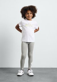 Camiseta blanca con el logo de "nike", leggins grises y zapatillas blancas. Niño de pie con las manos en las caderas, sonriendo, con el cabello rizado.