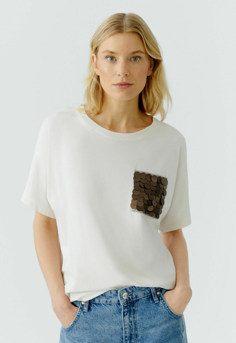 Witte T-shirt met korte mouwen en een ronde halslijn, met een donkerbruine pailletten zakdetail. Gekleed met lichtblauwe denim jeans.