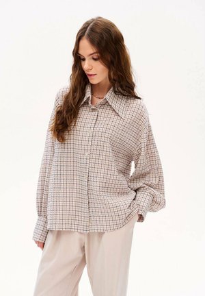 Młoda kobieta z długimi brązowymi włosami, ubrana w luźną, oversize'ową beżową koszulę w kratę na guziki oraz jasnobeżowe spodnie, spoglądająca w dół.