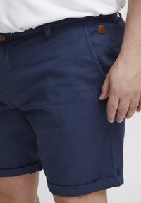 Marineblå shorts laget av lett materiale, med sidelommer, rullet kant og en brun skinnknapp som aksent på livet.