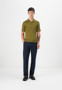 Polo de punto de manga corta en verde oliva con textura de canalé, combinado con pantalones azul marino. El modelo lleva mocasines negros, de pie sobre un fondo blanco.