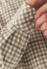 Cămașă în carouri maro și alb cu o textură moale; are manșete rotunde cu accente de nasturi din metal și un model distinctiv în carouri.