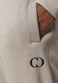 Gray cotton sweatpants feature a side pocket and a black embroidered logo on the left thigh, with a smooth, soft texture and ribbed waistband.