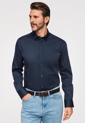 Homme barbu portant une chemise bleu foncé à manches longues boutonnée, un jean bleu clair et une ceinture marron foncé, avec une main dans la poche.