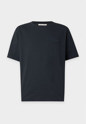 Mand iført en sort kortærmet T-shirt, lyseblå vidde jeans og brune sneakers, stående mod en ensfarvet hvid baggrund.
