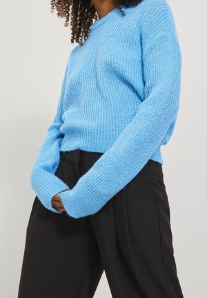 Person trägt einen hellblauen Strickpullover mit langen Ärmeln und schwarze, hoch taillierte Hosen, steht vor einem einfarbigen Hintergrund.