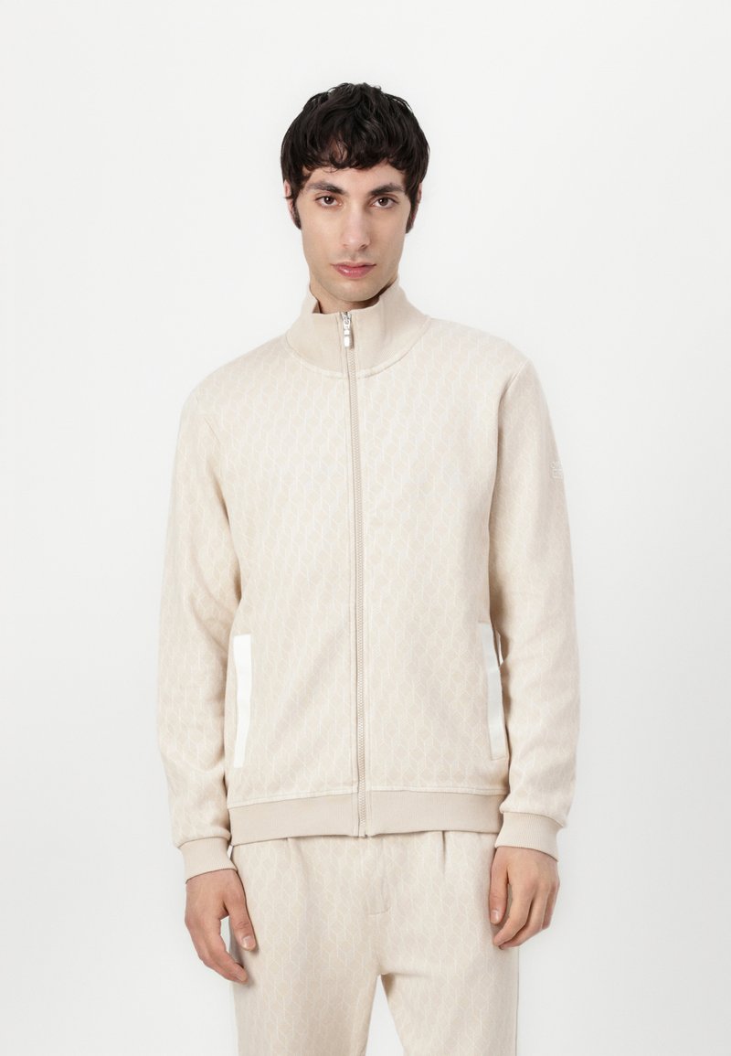 Veste beige zippée avec un motif en diamant subtil, col montant et poches latérales blanches. Tissu doux avec des poignets et un ourlet côtelés.