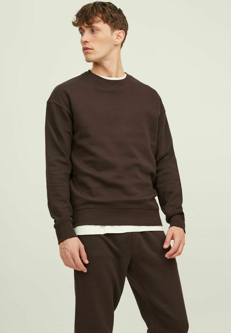 Bruin fleece sweatshirt met een ronde hals, zachte textuur en geribbelde boorden. Gedeeltelijk gedragen met een witte gelaagde shirt eronder.