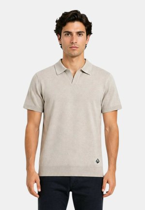 Giovane uomo con capelli scuri che indossa una polo beige a maniche corte e pantaloni scuri, in piedi davanti a uno sfondo neutro.