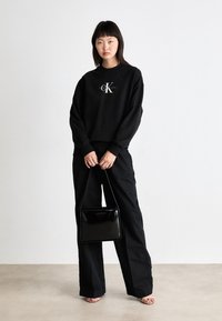Črn pulover z lahkim logotipom, v kombinaciji s črnimi širokimi hlačami. Model drži sijočo črno torbico v obliki kvadrata. Enostavna ozadja.