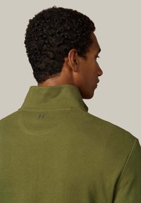 Polo de forro polar verde con un alto cuello, que presenta una tela texturizada y un sutil logo azul en la parte posterior del cuello.
