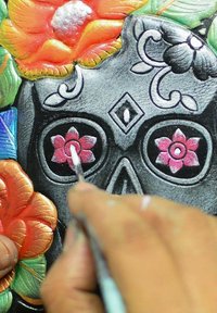 Schwarzer Schädel, verziert mit bunten Blumendesigns und komplexen Details. Eine Hand malt rosa Blumen auf die Augenhöhlen des Schädels.