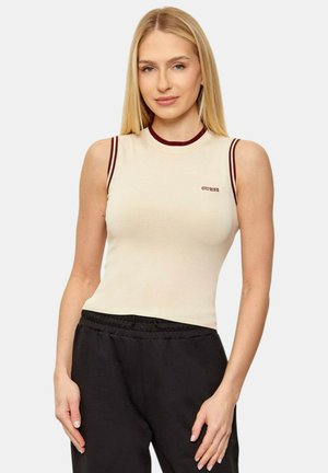 Donna bionda che indossa un top beige senza maniche con rifinitura color bordeaux e logo "GUESS", abbinato a pantaloni neri con vita elasticizzata, in piedi su sfondo bianco.