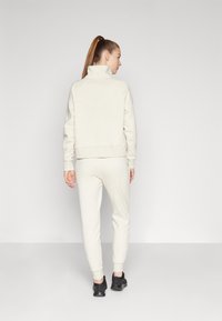 Hanorac și pantaloni de trening asortați, de culoare crem, dintr-un material moale, cu guler înalt și manșete ribbate. Pantofi sport negri completează ținuta.