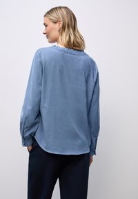 Camicia azzurro chiaro a maniche lunghe con colletto arricciato e polsini elastici. Il tessuto sembra morbido, con una texture liscia e una vestibilità comoda.