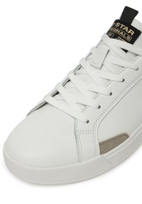 Biela kožená teniska s plochými bielymi šnúrkami, sivými semišovými prvkami, polstrovaným jazykom a čierno-zlatou logovou nášivkou na jazyku.