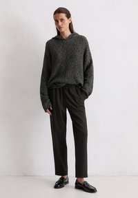 Haut à capuche en tricot gris, coupe oversize, avec un ourlet roulé, associé à un pantalon ample vert foncé et des mocassins noirs brillants.