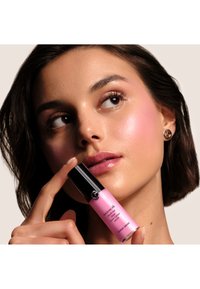 Transparent flaska med svart lock, som innehåller rosa vätska. Modell som håller flaskan nära ansiktet, med en jämn hy och markerade kindben.