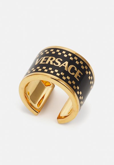 Anillo de puño en tono dorado con banda de esmalte negro, que presenta "VERSACE" en letras negritas y un patrón a cuadros a lo largo del borde.