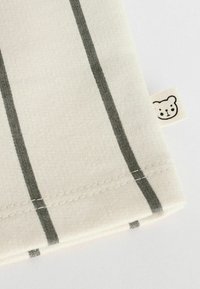 Tessuto di cotone con sfondo beige chiaro, caratterizzato da strisce verticali grigio scuro. Un piccolo etichetta con un grafiche di un orso è cucita sul bordo.