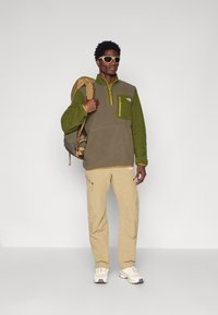 Pullover in pile verde oliva e grigio con tasca zip gialla; abbinato a pantaloni beige e scarpe da ginnastica bianche. Zaino sulla spalla.