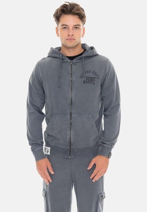 Jeune homme debout, portant un sweat à capuche gris zippé avec le texte « Boxing Dept Leone » et un pantalon gris assorti avec des poches latérales.