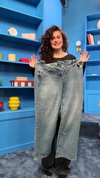 Γυναίκα με σγουρά μαλλιά χαμογελά, κρατώντας ένα υπερμεγέθες ξεθωριασμένο μπλε τζιν μπροστά από μπλε ράφια με πολύχρωμη διακόσμηση σε ένα μοντέρνο δωμάτιο.