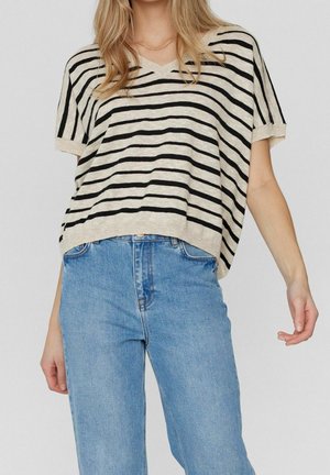 Vrouw die een loszittende beige en zwart gestreepte top met korte mouwen draagt, gecombineerd met lichtblauwe, hooggetailleerde jeans, staand tegen een effen achtergrond.