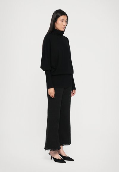 Melnā adīta turtlenecks džemperis ar plašām piedurknēm, kombinēts ar teksturētām melnām plašām biksēm ar saplēstiem malām un melniem augstpapēžu zābakiem.