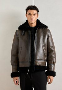 Giacca bomber in pelle marrone con colletto e polsini in shearling neri, caratterizzata da una cerniera frontale, tasche laterali e cuciture a contrasto.