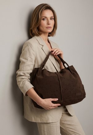 Vrouw in beige pak die een grote donkerbruine leren shopper vasthoudt, staande tegen een effen lichtgrijze achtergrond.