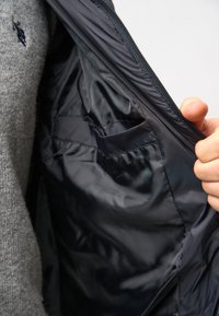 En hand som håller en öppen svart jacka med foder, som avslöjar en innerficka, bärs över en grå tröja.