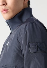 Jūras spēku zip-up jaka ar augstu apkakli, gludu tekstūru un izteiksmīgu melnu uzlīmi ar "Calvin Klein Jeans" un logotipu uz piedurknes.