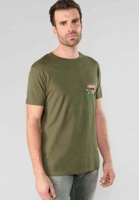 Hombre con cabello corto castaño y barba, vistiendo una camiseta de color verde oliva con un pequeño gráfico de un gato usando gafas de sol y la palabra "DOLLAR" en el pecho.