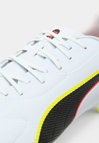 White athletic shoe with textured black sole, yellow and red accents, featuring flat laces and a streamlined design.