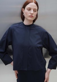 Navy cropped jacket with a high collar and wide sleeves. Smooth, lightweight fabric with subtle stitching details and side pockets.