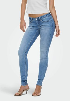 Kvinne iført lyseblå smale jeans, hvit avkortet topp, gjennomsiktige høyhælte sandaler og et armbånd på høyre håndledd.