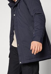 Mies, jolla on päällään laivastonsininen vetoketjullinen ja painonapeilla varustettu takki kermaisen valkoisen neuleen ja mustien housujen päällä, oikea käsi rentona sivulla.