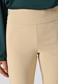 Primo piano di pantaloni beige a vita alta indossati con un top a maniche lunghe verde scuro, mostrando dettagli delle cuciture laterali e frontali.