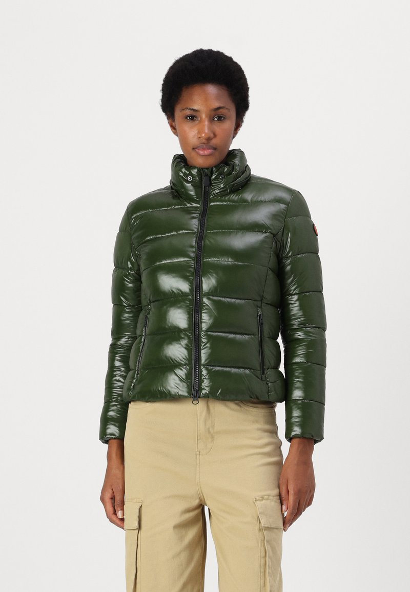 Groene donsjack met een glanzende afwerking, verticale gestepte ontwerp, hoge kraag, rits aan de voorkant en twee zijzakken met rits. Gdragend met beige broek.