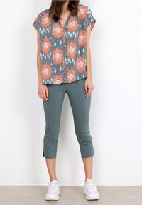 Kortærmet bluse med et farverigt blomsterprint i orange og teal, parret med ankel-lange, tætsiddende grønne bukser og hvide sneakers.