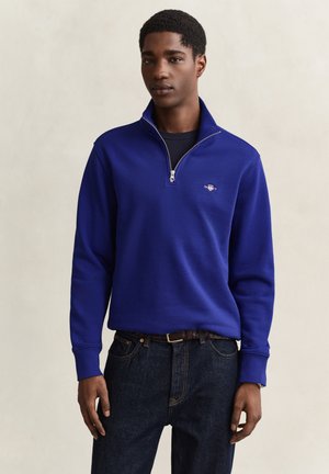 Jeune homme portant un pull bleu à fermeture éclair partielle sur une chemise noire et un jean foncé, debout devant un fond beige uni.