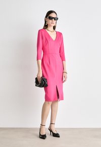 Fitted pink dress with a deep V-neck, three-quarter sleeves, and a front slit. Paired with black heels and a small black handbag.
