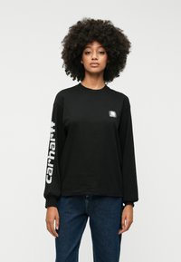 Melnas garām piedurknēm T-kreklas, izgatavots no kokvilnas, ar mazu logotipa uzlīmi krūšu daļā un spilgti baltu tekstu uz kreisās piedurknes.