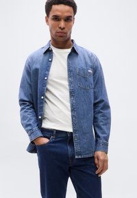 Πουκάμισο από τζιν σε ανοιχτό μπλε χρώμα, με κουμπιά και τσέπη στο στήθος. Φορεμένο πάνω από λευκό μπλουζάκι, συνδυασμένο με σκούρο μπλε τζιν παντελόνι. Λείο υφή.