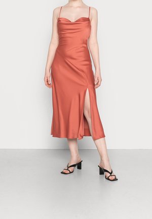 Copper satin slip dress with a cowl neckline, side slit, and adjustable spaghetti straps. Model wears black block-heel sandals.